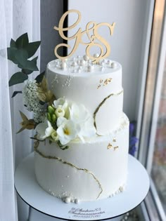 a three tiered cake with white flowers and gold initials on top is sitting in front of a window