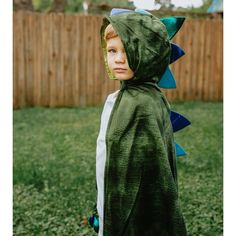 a young boy wearing a green dinosaur costume in the grass with a fence behind him