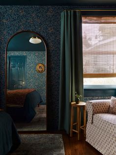 a bedroom with blue wallpaper and green drapes on the windowsill, a white chair in front of a mirror