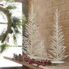 three white christmas trees in front of a brick wall with red berries on the table