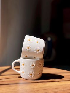 two white coffee cups sitting on top of a wooden table with daisies painted on them