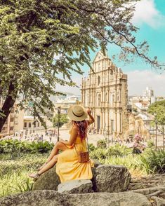 a woman sitting on top of a large rock next to a tree and some buildings