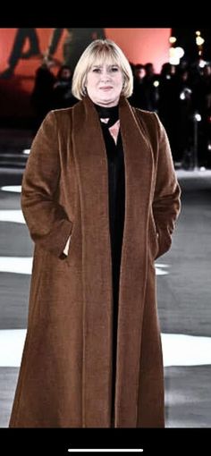 a woman in a brown coat is standing on the street with her hands behind her back