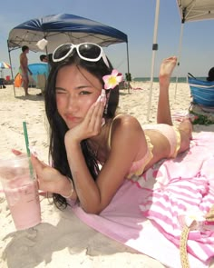 a woman laying on top of a beach next to a pink towel holding a drink