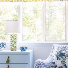 a blue and white chair sitting in front of a window next to a table with a lamp on it