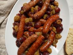 sausages and grapes on a white plate next to some breadsticks in the background