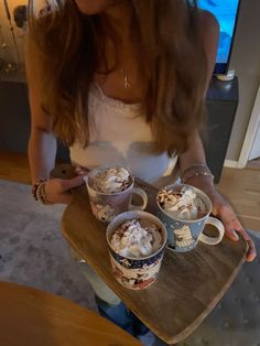 a woman holding two cups of hot chocolate and whipped cream on top of a wooden tray