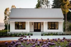 a white house with black doors and windows next to purple flowers in the foreground