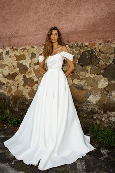 a woman standing in front of a stone wall wearing a white wedding dress with off the shoulder sleeves