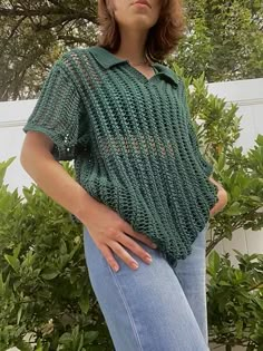 a woman standing in front of some bushes wearing a green knitted shirt and jeans