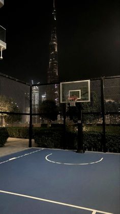 an empty basketball court in front of a tall building at night with the lights on