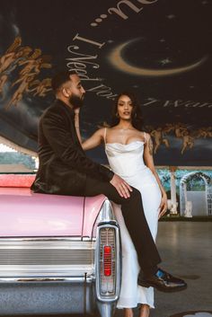 a man and woman sitting on top of a pink car in front of a sign