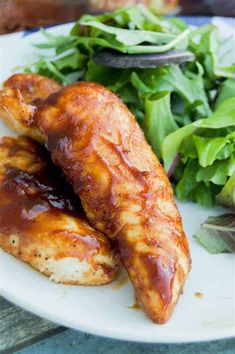 two pieces of chicken on a white plate next to a green leafy salad and an eggplant