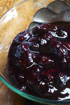 a glass bowl filled with blueberry sauce on top of a wooden table next to a metal spoon