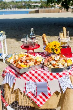 a picnic table with food and drinks on it at the beach near the water's edge