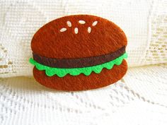 a felt hamburger brooch sitting on top of a white cloth covered table next to a lace doily