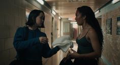 two women standing in a hallway talking to each other and one is holding a white mask