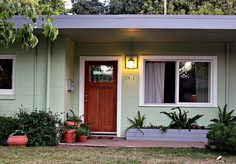 a small green house with two plants in front of it and a red door on the other side