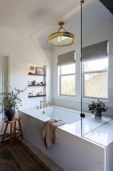 a white bath tub sitting under a window next to a wooden stool and potted plant