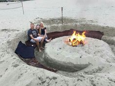 three people sitting around a fire pit on the beach with sand surrounding it and water in the background