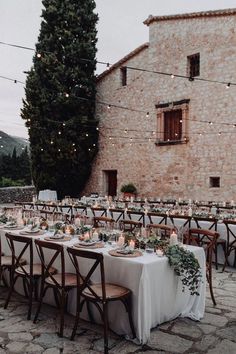 an outdoor dining table set up with candles and greenery on the tables in front of a stone building