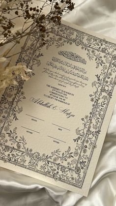 an ornate wedding card on a bed with white sheets and dried flowers in the foreground