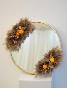 a round mirror sitting on top of a white pedestal next to a wall with dried flowers