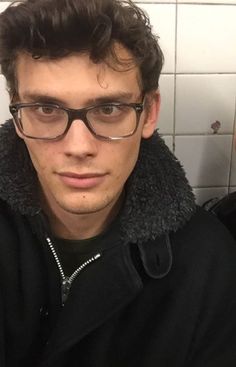 a man wearing glasses and a black jacket in a bathroom with white tiles on the wall