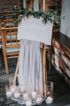 a wedding sign with candles and greenery on the back is surrounded by wooden chairs