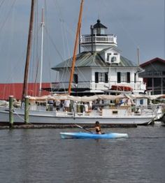 a person in a kayak paddling on the water near a light house and sailboats