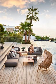 two people sitting on couches in front of a pool with palm trees and water