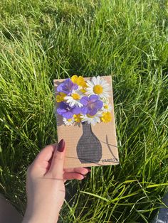 a person holding up a small card with flowers in the vase on it and grass behind them