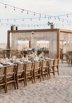 an outdoor dining set up on the beach with string lights strung over it and wooden chairs