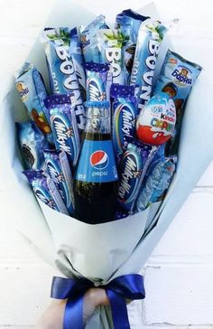 a hand holding a bouquet of chocolates and candy bar wrapped in blue ribbon with a white brick wall behind it