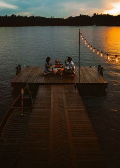 two people sitting on a dock with lights strung across the water and one person eating