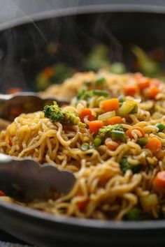 a wok filled with noodles and vegetables being stirred by tongs on the side