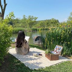 a woman sitting on a blanket in front of a pond reading a book and painting