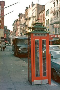 a red phone booth sitting on the side of a road next to parked cars and people