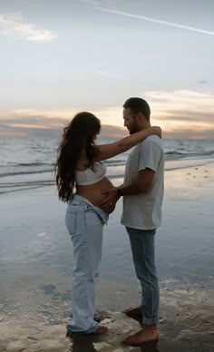 a pregnant woman standing next to a man on top of a beach near the ocean