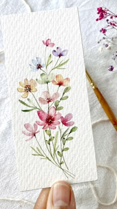 a person holding up a card with watercolor flowers on it and yarn in the background