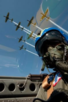 a man wearing a helmet and goggles looking out the window at airplanes flying in the sky