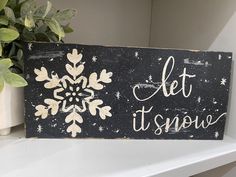 a wooden sign that says let it snow on top of a shelf next to a potted plant