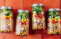 four mason jars filled with different types of food on a red brick wall, each containing beans and vegetables