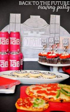 a table topped with pizza and cupcakes next to cans of soda