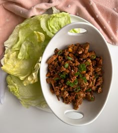 a white bowl filled with meat and lettuce on top of a table next to a pink cloth