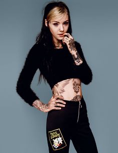 a woman with tattoos on her body posing in black pants and cropped top, leaning against a blue wall