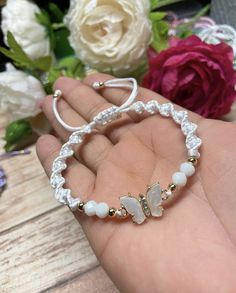 a hand holding a bracelet with white beads and butterfly charms on it next to flowers