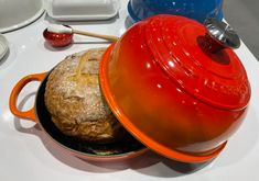 an orange pot with a red lid next to a loaf of bread on a white table