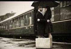 a man and woman standing next to each other in front of a train with an umbrella