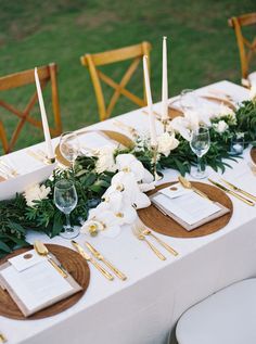 the table is set with white flowers and greenery, gold place settings and candles